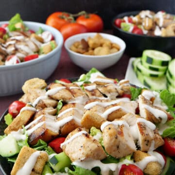 Two black bowls and a white bowl filled with salad, drizzled in ranch and a plate of sliced cucumbers in the background.