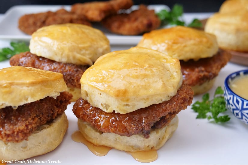 A horizontal photo of a white surface with four honey butter chicken biscuits on it with fried chicken in the background on a white plate.