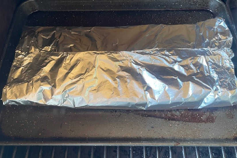 A baking sheet with a foil pack of chicken strips wrapped up and on the smoker.
