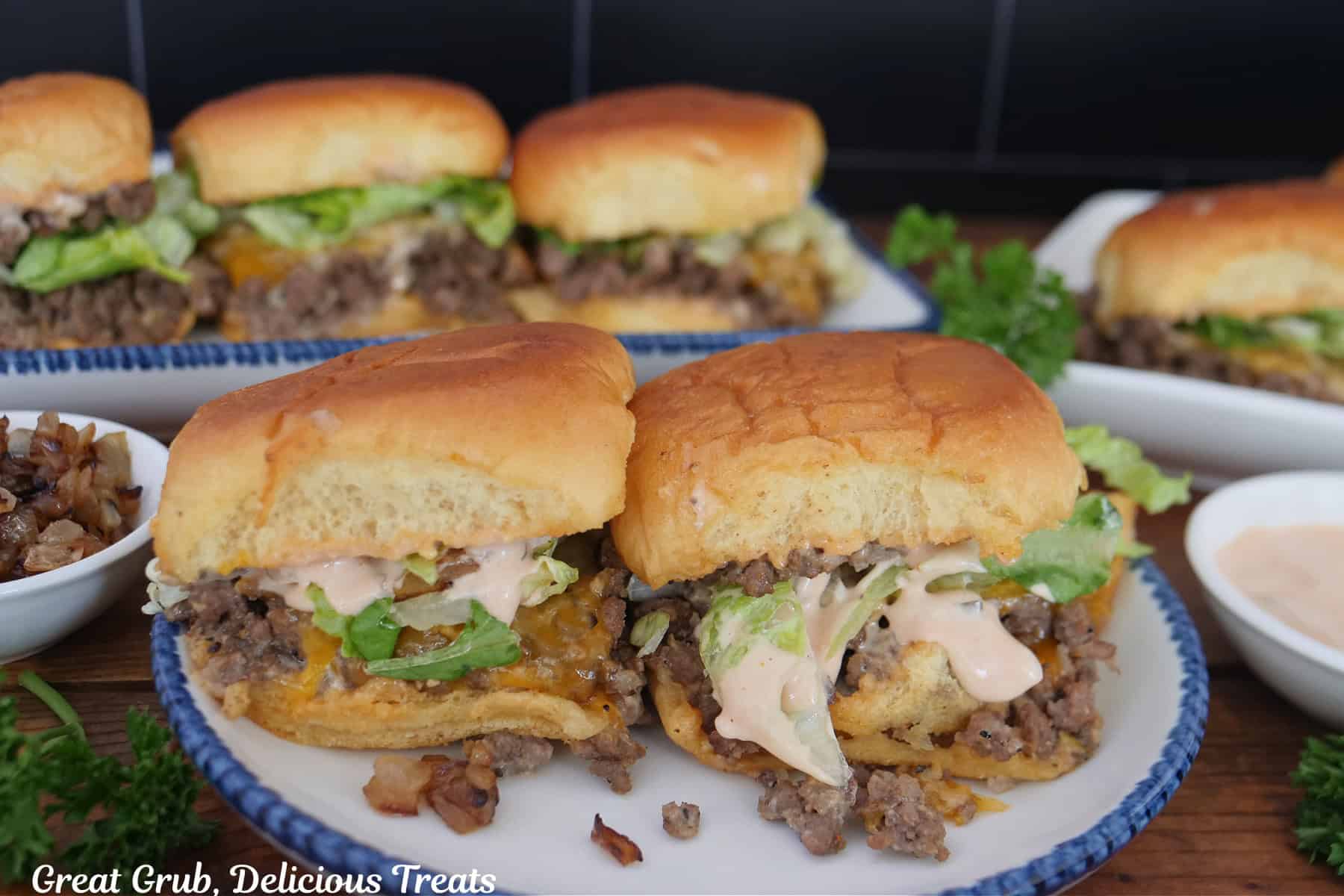 Two beef sliders on a white plate with blue trim with more sliders in the background.