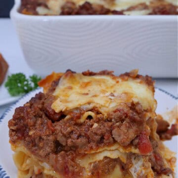 A round white plate with blue trim with a serving of lasagna bolognese on it.
