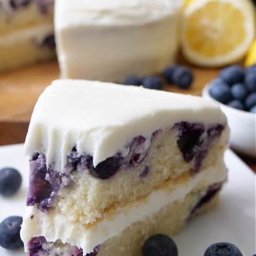 A slice of lemon blueberry cake on a white plate with lemon cream cheese frosting.