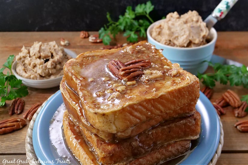 Maple Pecan French Toast is a homemade french toast recipe made with thick Texas toast, finely chopped pecans, cooked until golden brown, topped with delicious maple pecan butter and maple syrup. Four pieces of French toast stacked up on a blue plate with syrup and pecans on top, and two small bowls of whipped butter in the background.