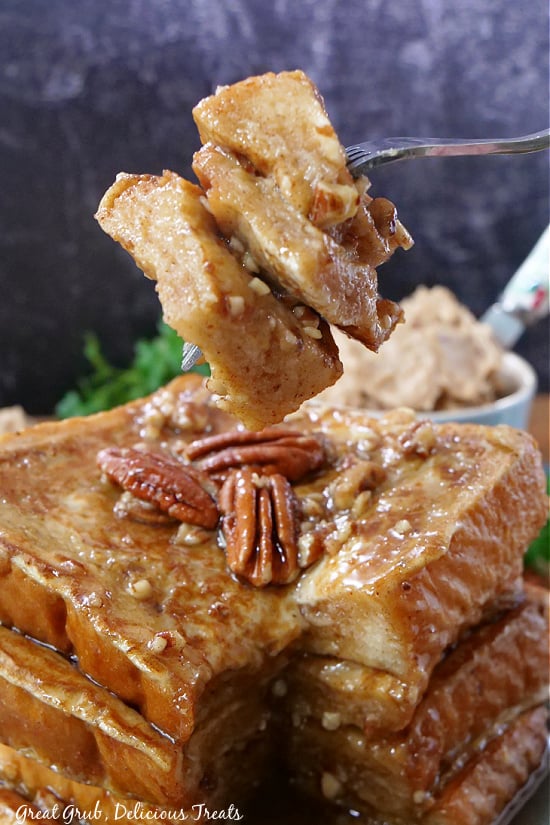 Maple Pecan French Toast is the perfect breakfast recipe; thick Texas toast, coated in cinnamon and finely chopped pecans, cooked until golden brown, and topped with a whipped maple butter, and maple syrup. A bite of French toast on a fork, with a stack of French toast in the background.