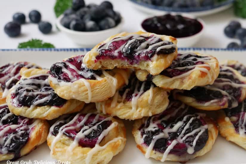 A horizontal photo of a white plate with blue trim filled with mini blueberry pastries pile on it and the top pastry has a bite taken out of it.