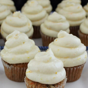 A white surface with mini carrot cake cupcakes on it with orange buttercream frosting on each cupcake.