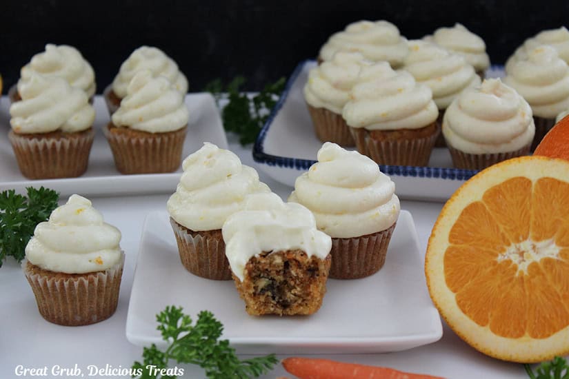 A white surface with white plates filled with mini carrot cake cupcakes with frosting on them.