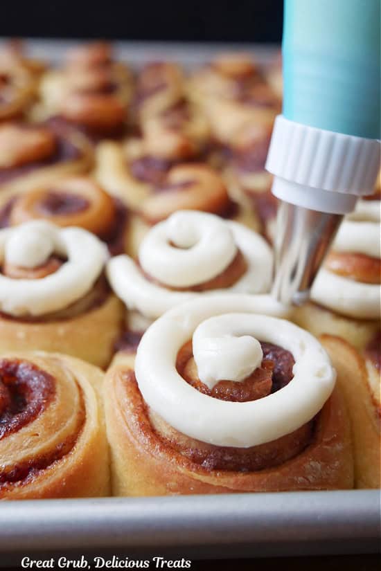 Mini cinnamon rolls on a baking sheet with cream cheese icing being swirled on top of them.