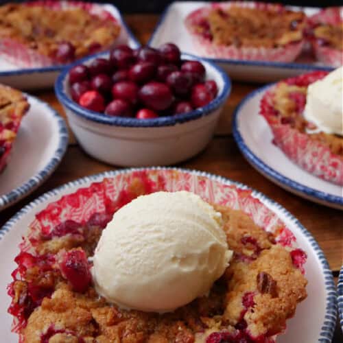 A wood surface with white plates with blue trim with a mini cranberry cobbler on them and a bowl filled with fresh cranberries.
