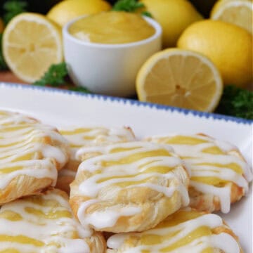 A white plate with blue trim with 6 mini lemon pastries on it with lemons in the background.