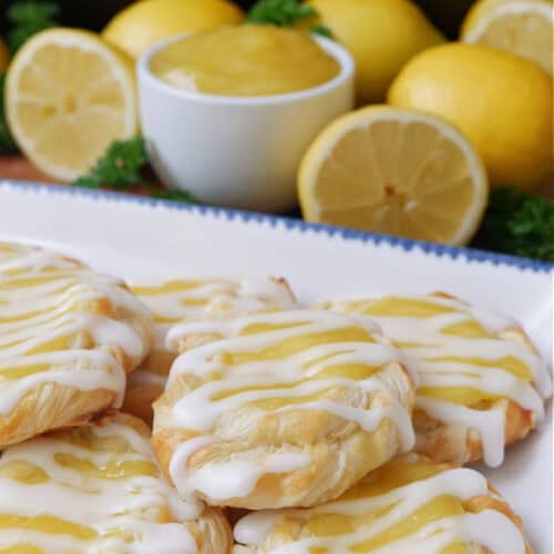 A white plate with blue trim with 6 mini lemon pastries on it with lemons in the background.