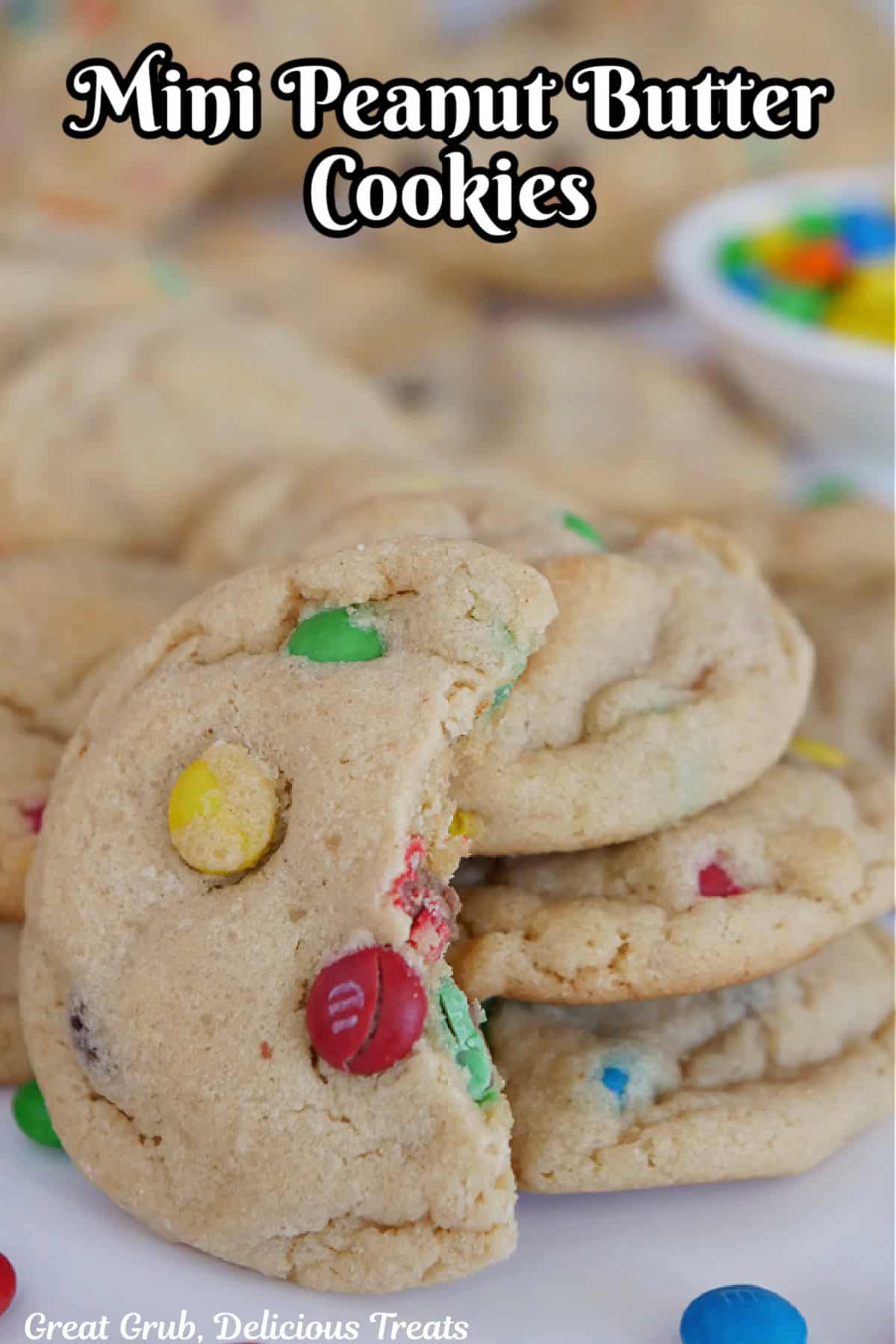 A white surface with mini peanut butter cookies placed on the surface with a few stacks of cookies and one with a bite taken out of it.