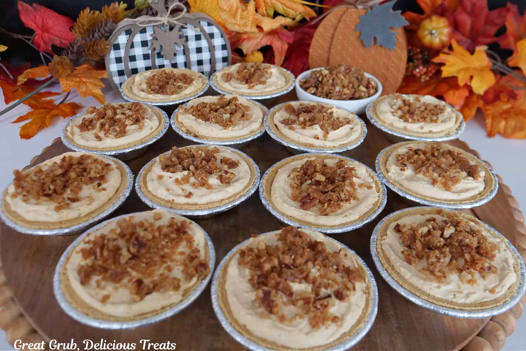 A wood surface with twelve mini pumpkin pies on it with fall foliage in the background.