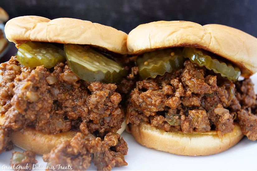 Two sloppy joes on a counter with pickles on top of the meat.