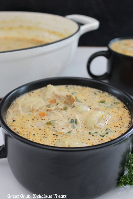 A black soup bowl with two handles filled with chicken gnocchi soup and a white pot in the background filled with soup.