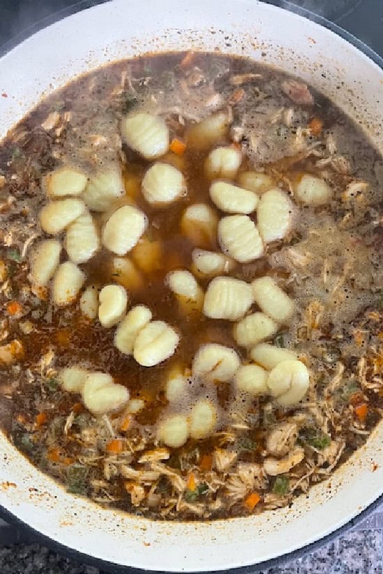 A white pot with shredded chicken, veggies, and potato gnocchi in a broth.