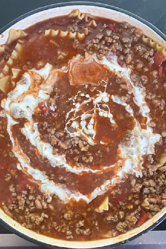 A white pot with all the ingredients for lasagna soup in it and heavy cream added.