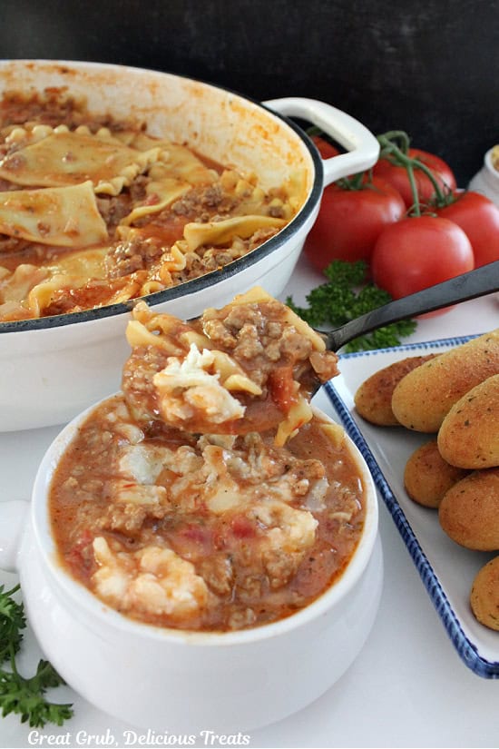 A white surface with a white soup bowl filled with a serving of lasagna soup with the pot of soup in the background and a plate with bread sticks on it off to the side.