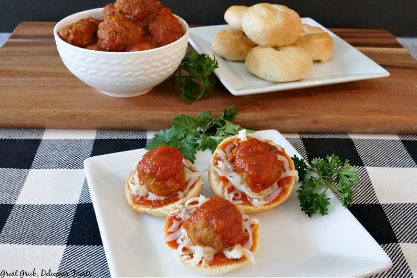 A picture of Meatball Bagel Bites on a white plate with parsley, cooked meatballs in a white bowl, and plain bagels on a white plate in the background on a brown cutting board.