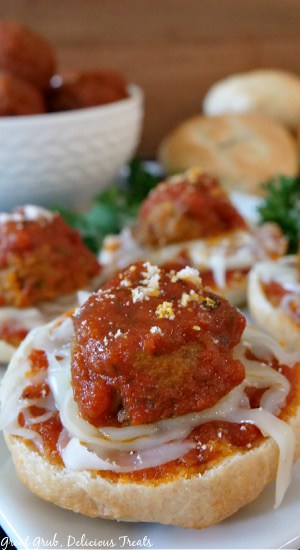 Meatball Bagel Bites are loaded with delicious flavor and are a delicious appetizer that the whole family will enjoy. A close up picture of a Meatball Bagel Bite on a white plate with bagel bites in the background, along with parsley, cooked meatballs covered in spaghetti sauce in a white bowl, and plain bagels.