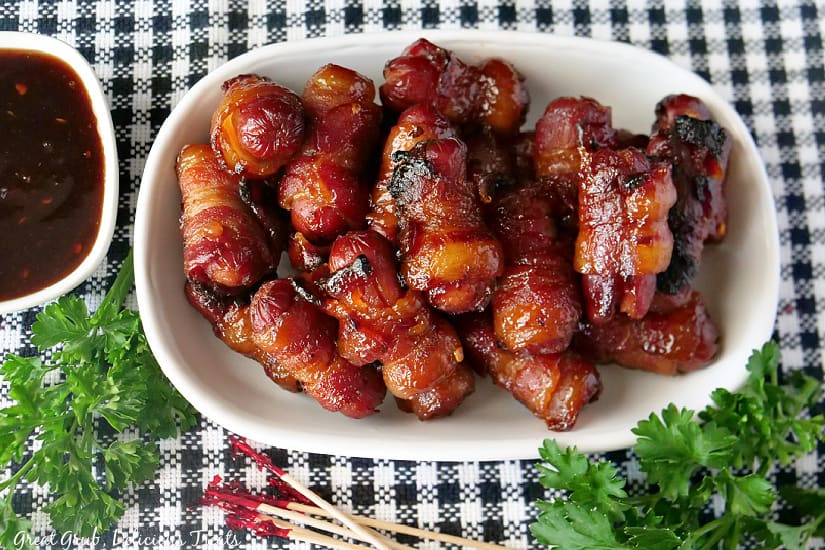 A black and white placemat with a white oval bowl filled with little smokies wrapped in bacon with a small bowl filled with barbecue sauce. placed on it.