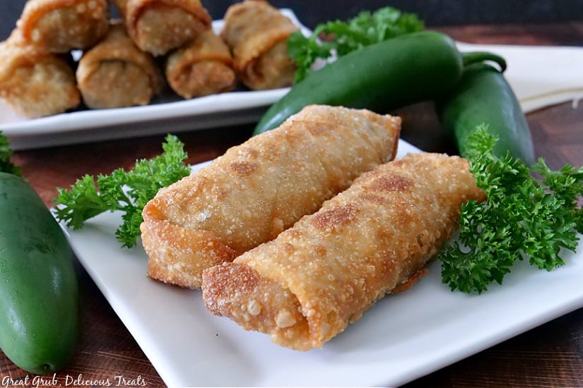 Cheesy Ground Beef Egg Rolls are the perfect appetizer recipe, loaded with ground beef, mozzarella and cheddar cheese, seasoned perfectly and fried to perfection. 2 Cheesy Ground Beef Egg Rolls on a white plate with another white plate in the background with 6 more egg rolls on it.