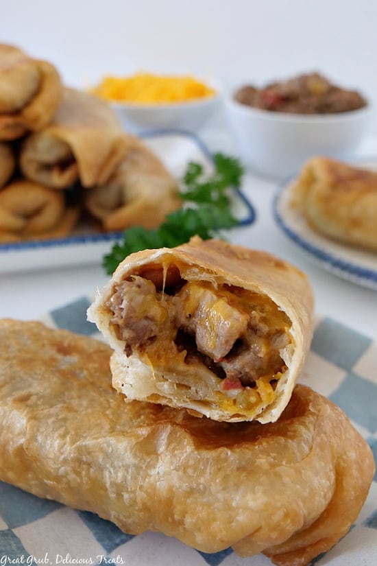 Fried Burritos are packed full of deliciously seasoned beef, pork, ground beef, two types of cheese, beans and more. Two fried burritos on a white and blue checkered plate with one burrito showing the inside filling of the burrito.