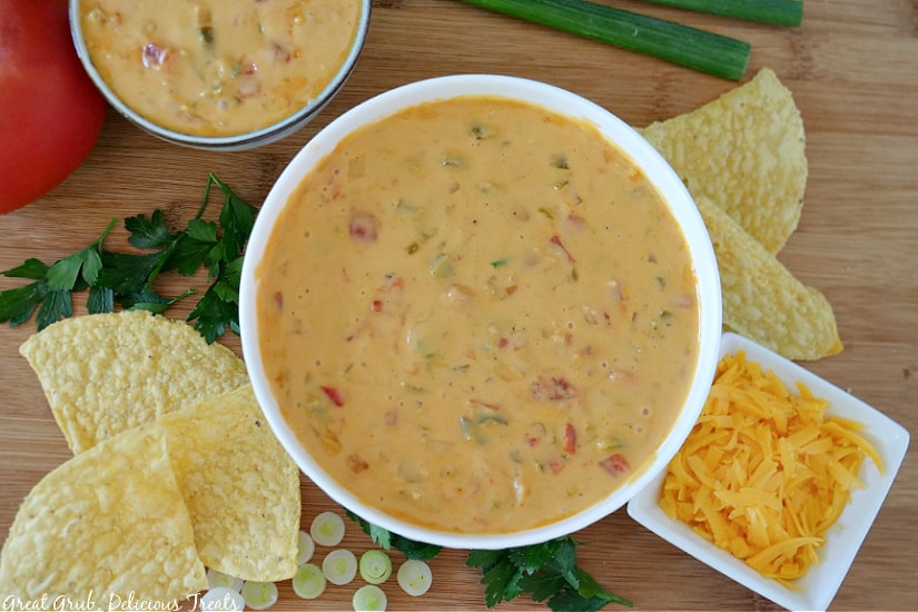 Spicy Queso is an easy to make, delicious dip recipe packed with flavor. Two white bowls filled with spicy queso sitting on a wood cutting board with tortilla chips, shredded cheese, cilantro and sliced green onions along side of the bowls.