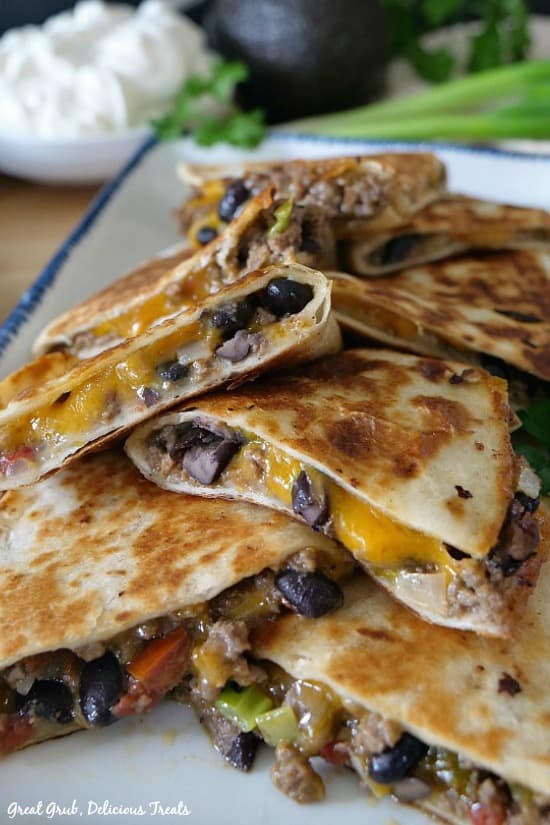 Ground Beef Quesadillas are filled with ground beef, black beans, two types of cheese and fried to a golden brown. A white rectangle plate with blue trim with eight quesadilla wedges layered over each other.