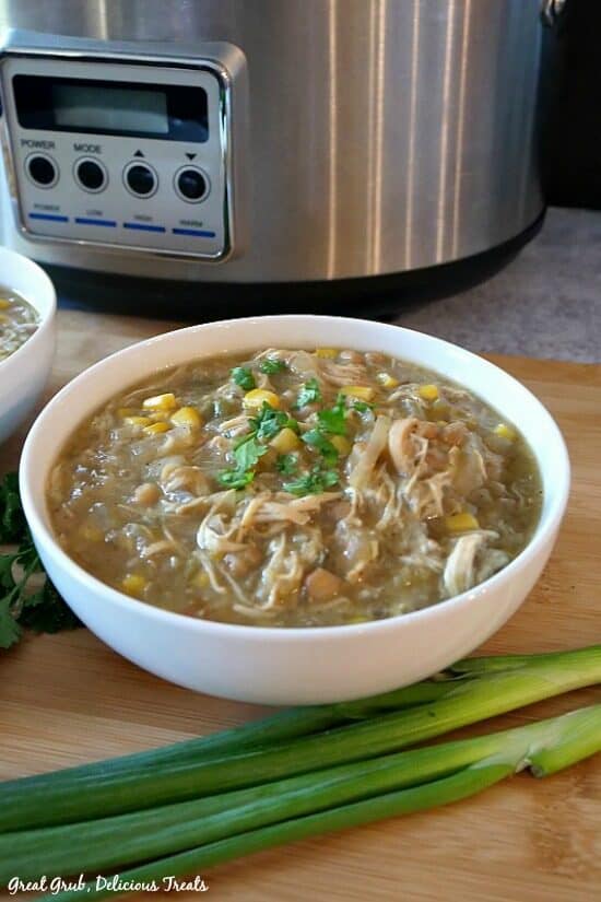Slow Cooker White Chicken Soup is loaded with chicken, beans, corn and more, slow cooked and makes an easy dinner idea. White bowl with white chicken chili in it, green onions in the foreground, and crockpot in the background