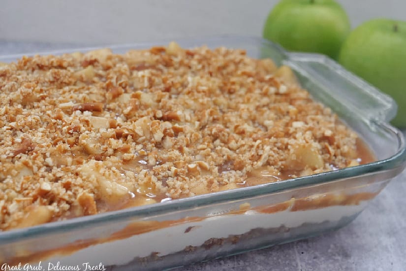 Apple Cinnamon Pretzel Salad is an easy, delicious pretzel salad recipe. A 9 x 13 baking dish with apple cinnamon pretzel salad in it with two green apples in the background.