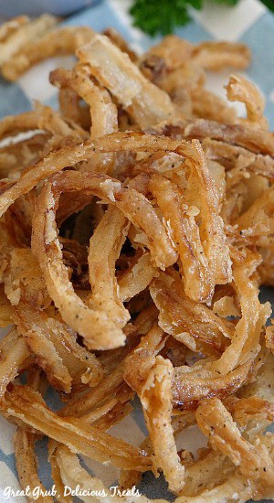 Fried Onion Strings are fried to a crispy crunch and loaded with flavor and make a delicious appetizer or snack recipe. Close up picture of Fried Onion Strings on a white and blue checkered plate.