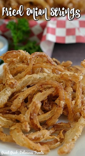 Fried Onion Strings are a delicious onion recipe that is simple to make, are crispy, crunchy, and full of flavor. Fried Onion Strings on a white plate with a basket filled with onion rings in the background.