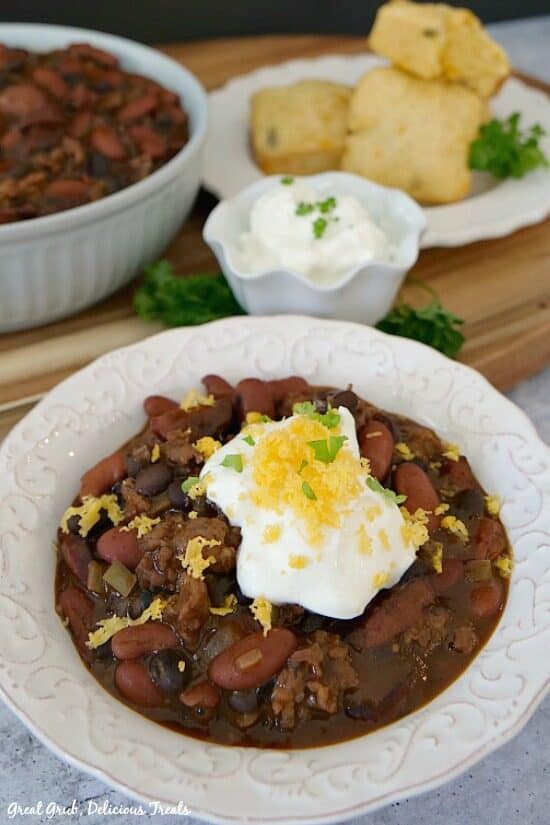 One pot kielbasa chili is a deliciously filling and flavorful one pot dinner. 2 white bowls full of chili with sour cream and grated cheese on top, a small white bowl with a dollop of sour cream, and a white plate with pieces of cornbread.