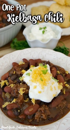 One Pot Kielbasa Chili is a delicious, flavorful one pot chili recipe loaded with kielbasa, onions, and bell peppers. A picture of One Pot Kielbasa Chili in a white bowl topped with sour cream and shredded cheese with a white bowl full of chili, a small white bowl of sour cream, and a white plate with cornbread.