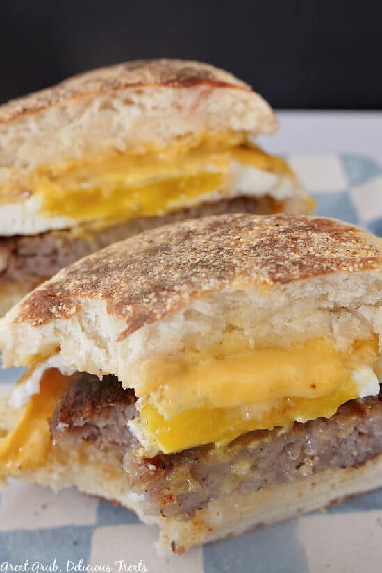 Sausage and Egg Breakfast Sandwiches are delicious sandwiches that are filled with protein and are the perfect way to start the day. A breakfast sandwich cut in half, on a white and blue checkered plate, with a bite taken out of it.