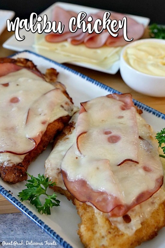 Two pieces of breaded and fried chicken breasts, on a white plate with blue trim, with melted cheese and ham on top with extra slices of ham and Swiss cheese and dipping sauce in the background.