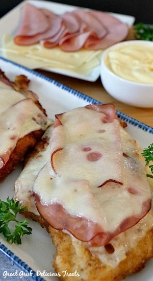 Malibu Chicken is a delicious fried chicken recipe that is topped with ham and melted swiss cheese and is delicious dipped in a homemade mayonnaise and mustard sauce. Malibu Chicken on a white plate with sliced ham and cheese in the background on a white plate with a dipping sauce in a white bowl.