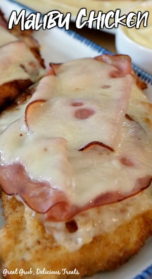 Malibu Chicken is a delicious chicken recipe that is topped with ham and melted swiss cheese, paired with a delicious mayonnaise-mustard sauce. A breaded chicken breast that is fried and topped with Swiss cheese and ham on a white plate with blue trim.