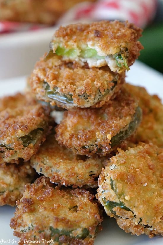 Deep Fried Crispy Jalapenos are crispy, crunchy and deliciously seasoned sliced jalapenos, fried until golden brown. A pile of Deep Fried Crispy Jalapenos stacked up on a white plate with one of the jalapenos with a bite taken out of it showing the sliced jalapeno surrounded by the crunchy coating.