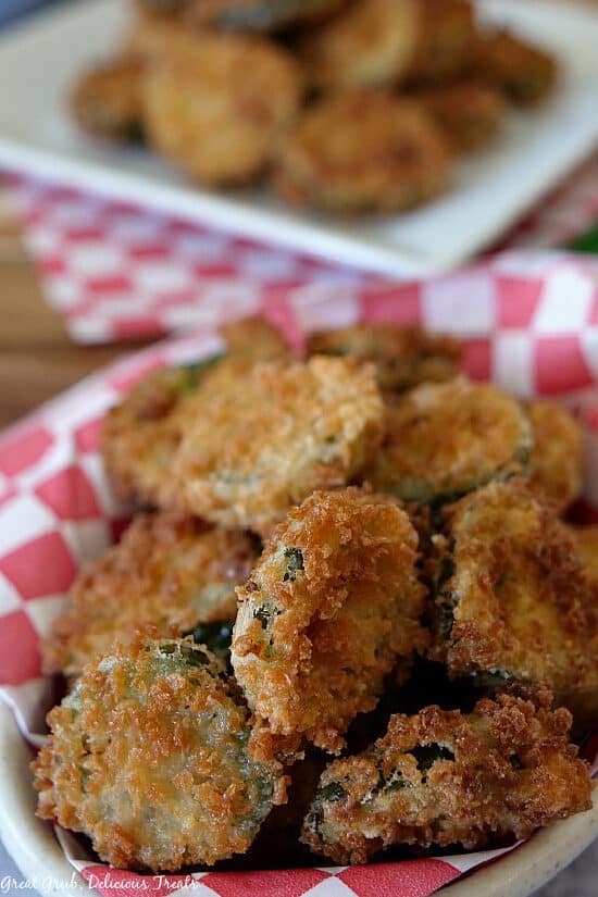 Deep Fried Crispy Jalapenos are seasoned perfectly, fried until golden brown and crispy and are a favorite snack recipe everyone will enjoy. Deep Fried Crispy Jalapenos are in a bowl lined with red and white checked paper with more fried jalapenos in the background on a white plate.