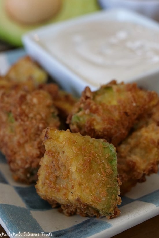 Crispy Fried Avocado Bites are a delicious bite-size appetizer recipe perfect for fame day parties, family gatherings or as a tasty snack. A small pile of crispy fried avocado bites on a light blue and white checkered plate with a small white square bowl filled with ranch dressing for dipping on the plate as well.