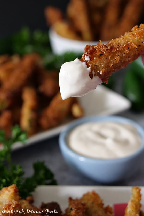Crispy Jalapeno Fries are a delicious appetizer recipe with a crunchy coating and a thin strip of jalapeno on the inside. A photo of a crispy jalapeno fry being held up after being dipped in ranch dressing with more jalapeno fries in the background on a plate and in a white bowl.