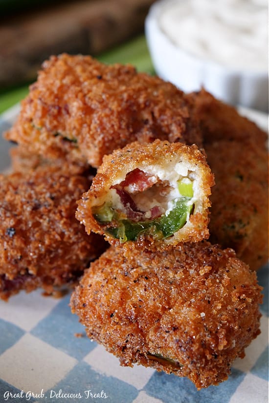Stuffed Jalapeno Poppers are the perfect appetizer recipe, stuffed with a delicious cream cheese mixture consisting of bacon, green onions, cheese and seasoned deliciously. A close up view of a stack of stuffed jalapeno poppers with a bite taken out of one showing the jalapeno, cream cheese mixture and bacon all sitting on a blue and white checker plate.