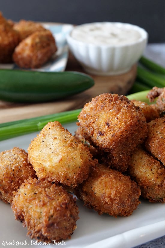 Stuffed Jalapeno Poppers are loaded with flavor, stuffed with cream cheese, bacon, cheese, green onions and fried to perfection. A stack of Jalapeno poppers on a white plate with a jalapeno and green onions, more poppers and a white bowl filled with ranch dressing in the background.