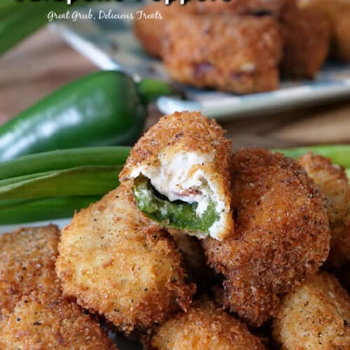 Stuffed jalapeno poppers stacked up on a white plate with a bite taken out of one of them, with green onions and a jalapeno in the background, and another white plate with jalapeno poppers stacked up.