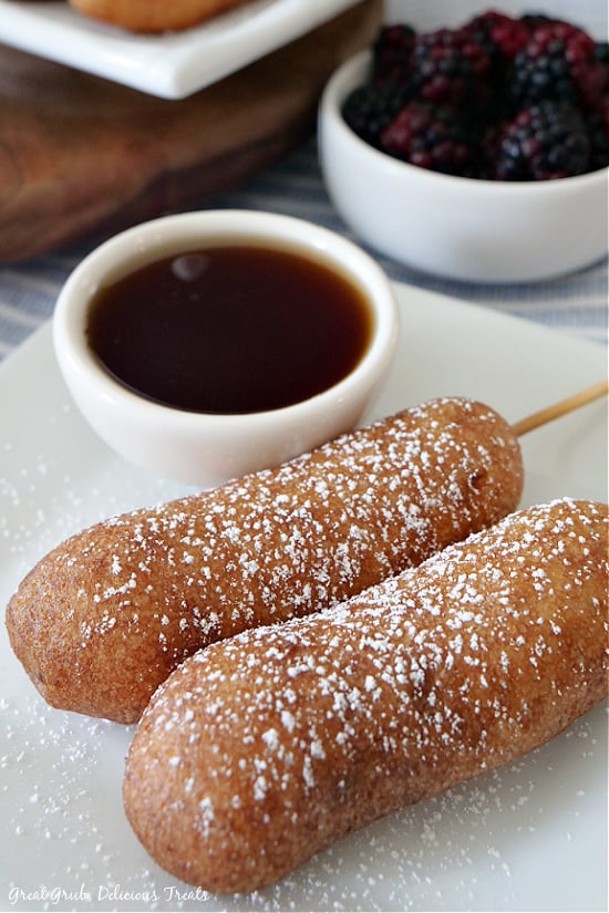 Pancake Sausage on a Stick are easy to make, delicious and a favorite pancake recipe. Two pancake sausages on a stick placed on a white plate with a small white bowl filled with maple syrup and another white bowl filled with fresh blackberries.