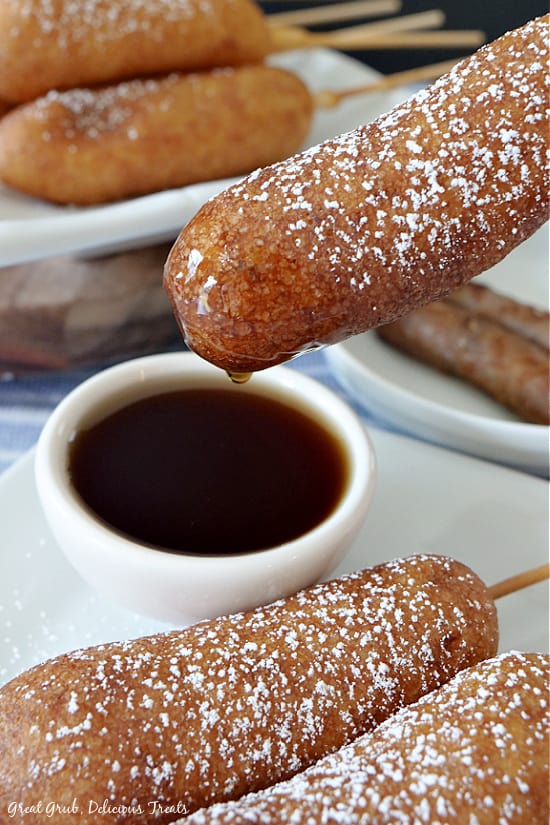 Pancake Sausage on a Stick is an easy, delicious, pancake recipe with a cooked sausage in the middle, then deep fried until golden brown. a Pancake Sausage on a Stick being dipped into maple syrup.