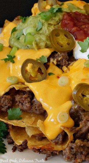 Mile High Nachos is a delicious nacho recipe that is loaded with ground beef, refried beans, shredded cheese, and nacho cheese sauce, topped with jalapenos, green onions, cilantro, homemade guacamole, salsa, and sour cream. A super close up of nachos on a white plate.