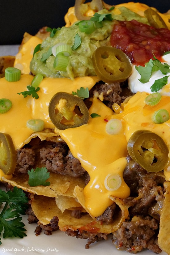 Mile High Nachos are a delicious recipe that is loaded with ground beef, refried beans, nacho cheese sauce, salsa, fresh guacamole, jalapenos, green onions, and cilantro. A close up picture of Mile High Nachos stacked up, showing ground beef, nacho cheese, green onion, guacamole, salsa. sour cream, jalapenos, and cilantro.
